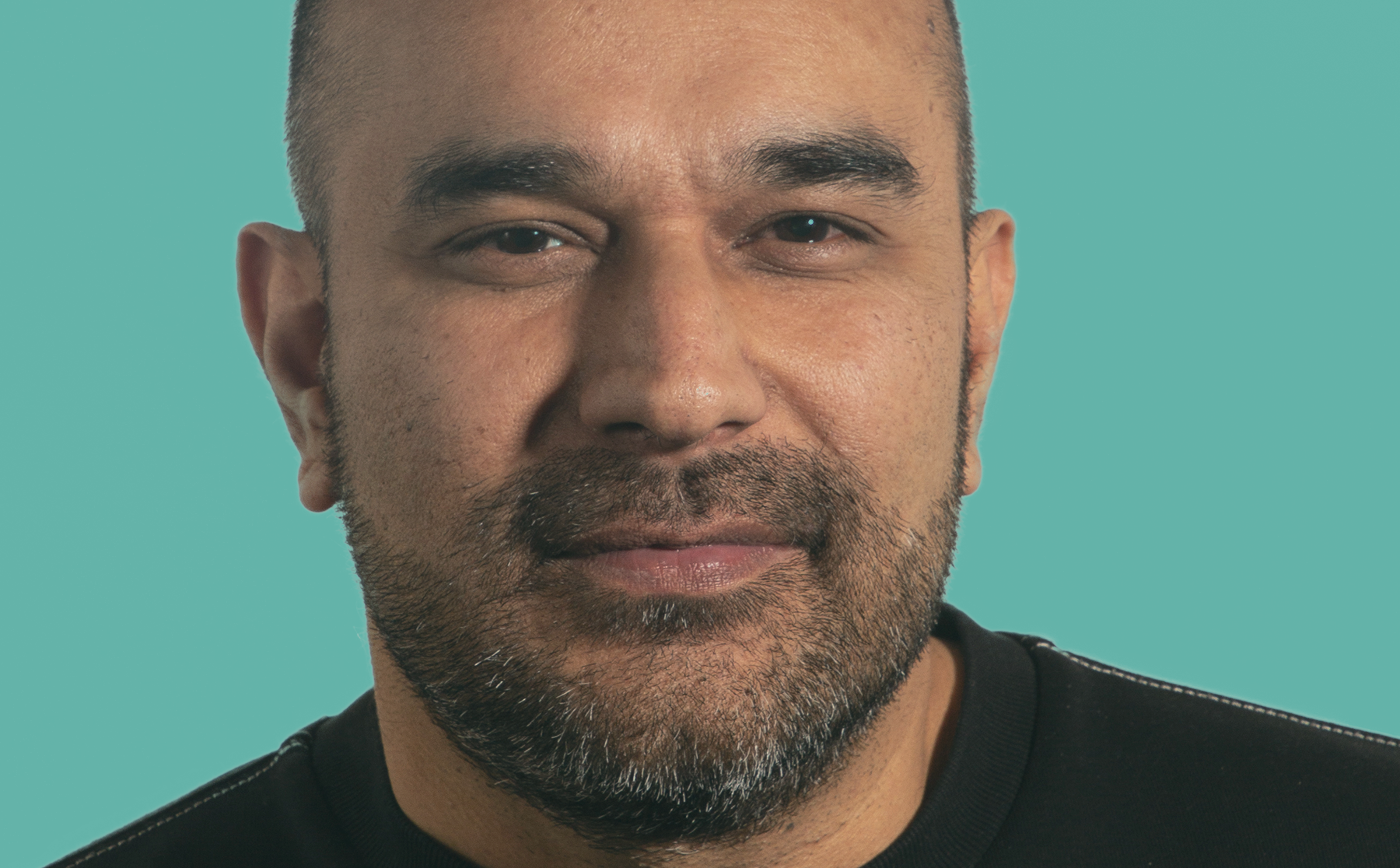 An asian man with a beard is wearing a dark top and standing in front of a blue background.